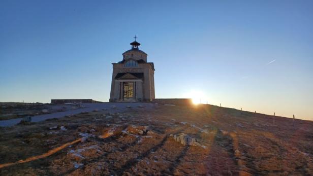 Kleine Kapelle auf einem Hügel bei Sonnenaufgang, mit klarem Himmel und spärlicher Vegetation im Vordergrund.