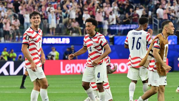 Mehrere Fußballspieler der US-Nationalmannschaft jubeln lachend auf dem Spielfeld, während Fans im Hintergrund feiern.