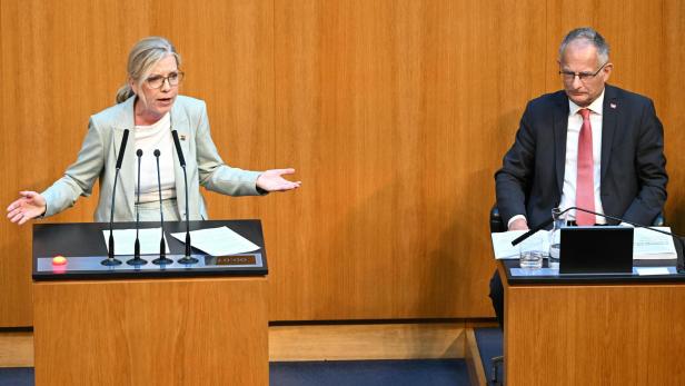 Eine Frau spricht gestikulierend an einem Rednerpult, während ein Mann daneben sitzt und nach unten blickt.