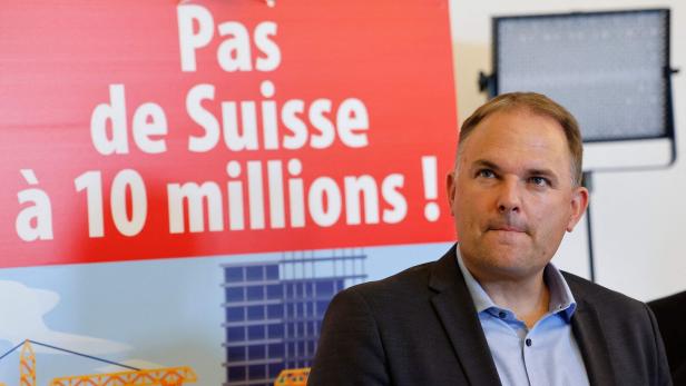 Ein Mann im Anzug steht vor einem roten Plakat mit der Aufschrift „Pas de Suisse à 10 millions!“.