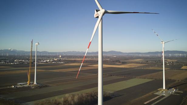 Mehrere Windkraftanlagen stehen auf weiten Feldern, eine wird gerade mit einem Kran aufgebaut, im Hintergrund Berge.