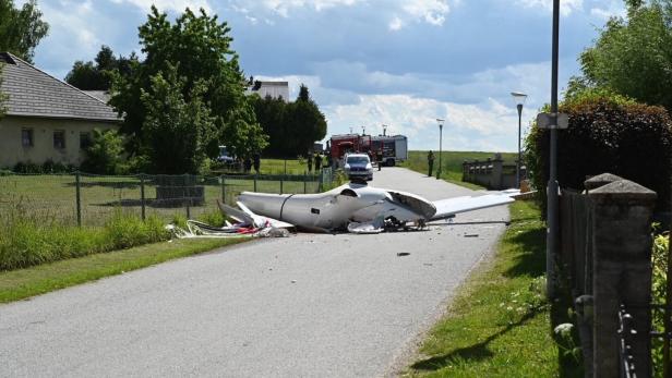 Ein abgestürztes Kleinflugzeug liegt zerstört auf einer Landstraße, Einsatzfahrzeuge und Einsatzkräfte im Hintergrund.