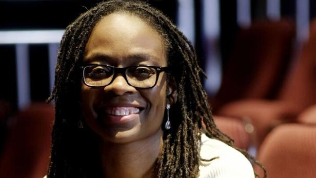 Eine Frau mit Brille und langen Dreadlocks sitzt lächelnd auf einem gepolsterten Stuhl in einem Saal mit roten Sitzreihen.