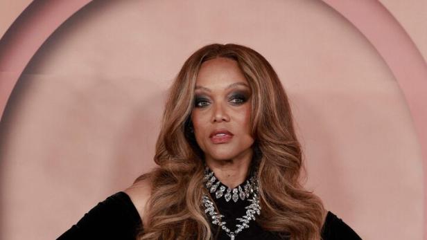 Tyra Banks trägt ein schwarzes, schulterfreies Kleid mit silbernen Verzierungen, langen Handschuhen und einer auffälligen Halskette vor einem Bogen.