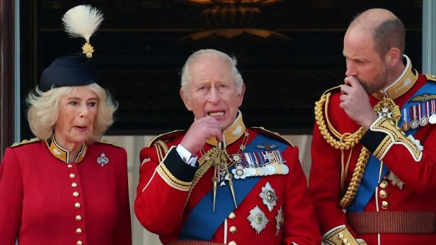 König Charles III., Königin Camilla und Prinz William in prunkvollen roten Uniformen auf einem Balkon.