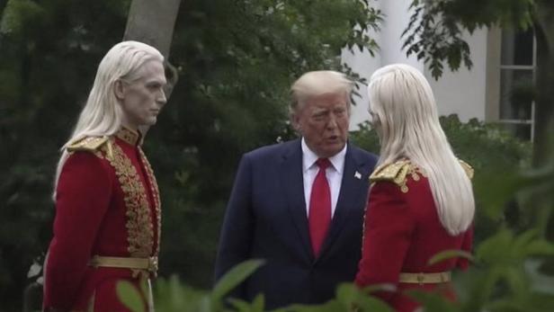Donald Trump steht im Garten des Weißen Hauses zwischen zwei Personen mit sehr blasser Haut, weißblondem Haar und roten Uniformen mit goldenen Verzierungen.