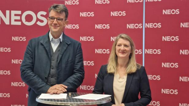 Zwei Personen stehen an einem Stehtisch vor einer pinken Wand mit mehrfachem NEOS-Schriftzug.