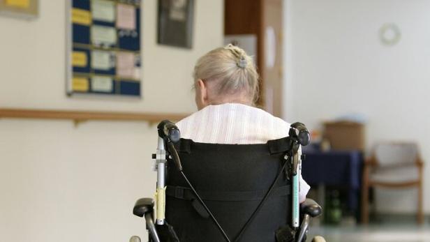 Eine ältere Person mit grauen Haaren sitzt im Rollstuhl auf einem Flur mit hellen Wänden und einigen Stühlen.