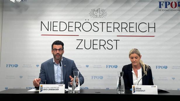 Zwei Personen sitzen an einem Tisch mit Namensschildern vor einer Wand mit der Aufschrift „Niederösterreich zuerst“.