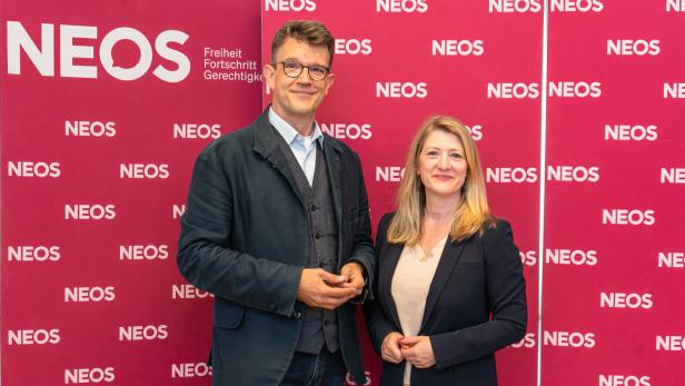 Ein Mann und eine Frau stehen lächelnd vor einer roten Wand mit dem wiederholten Logo der Partei NEOS.