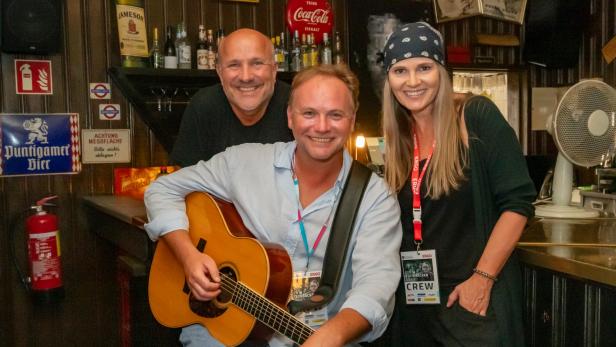 Drei fröhliche Menschen in einer Bar, einer hält eine Gitarre, die anderen stehen daneben und lächeln.