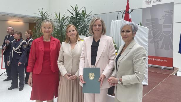 Vier Frauen in eleganter Kleidung stehen lächelnd nebeneinander, eine hält eine Urkunde mit österreichischem Wappen.