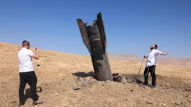 Zwei Männer in einer Wüstenlandschaft neben einem im Boden steckenden Raketenwrack, wobei einer den Vorfall fotografiert.