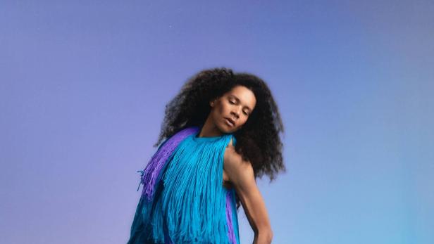 Rebecca Horner trägt ein fließendes Fadenkleid von Franz Josef Baur vor blauem Studiohintergrund.