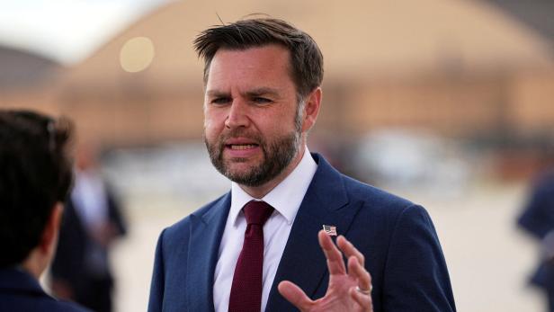 J.D. Vance spricht in einem dunkelblauen Anzug mit roter Krawatte und einer kleinen US-Flaggen-Anstecknadel gestikulierend zu einer Person.
