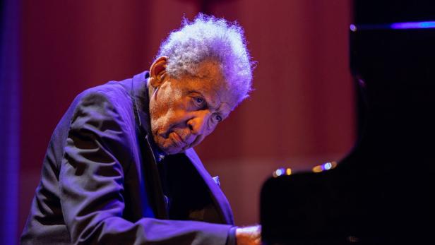 Ein älterer Mann spielt konzentriert Klavier auf einer Bühne, violett beleuchtet.
