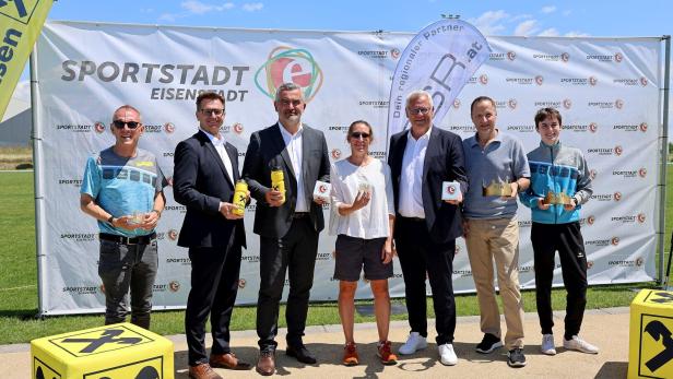 Sechs Erwachsene und ein Jugendlicher posieren vor einer Sportstadt-Eisenstadt-Wand, teils mit Trinkflaschen und kleinen Trophäen.