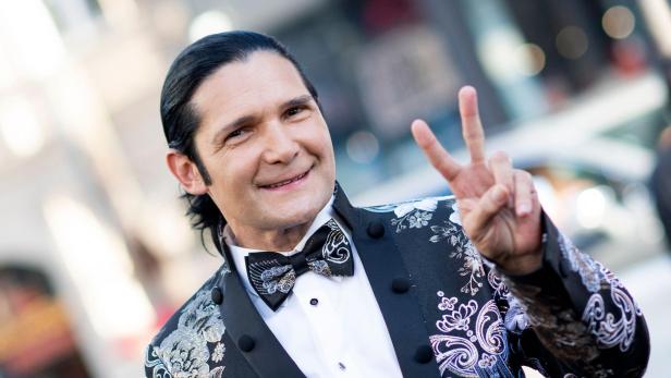 Corey Feldman lächelt in einem gemusterten Sakko mit Fliege und zeigt ein Friedenszeichen.