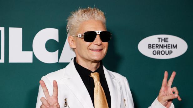 Corey Feldman mit blonden Haaren und Sonnenbrille trägt einen weißen Anzug vor einem Hintergrund mit dem Logo der The Bernheim Group.