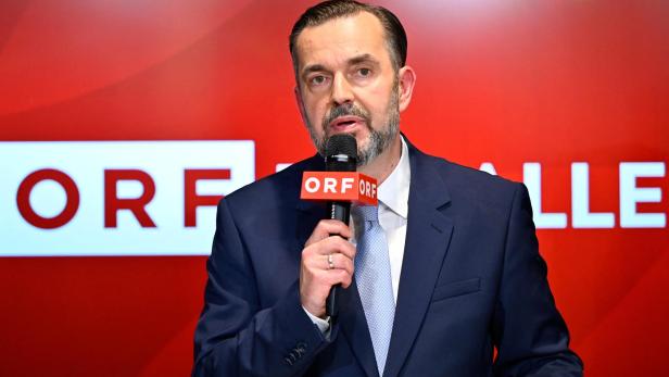 Mann im Anzug spricht in ein ORF-Mikrofon vor rotem Hintergrund.