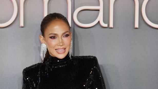 Jennifer Lopez trägt ein schwarzes Paillettenkleid mit einer großen pinken Schleife vor einem grauen Hintergrund.