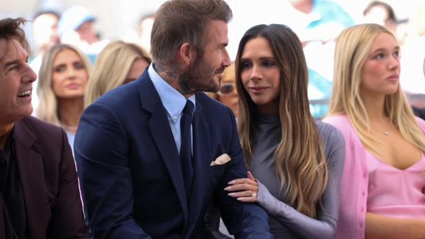 David Beckham im Anzug und Victoria Beckham in einem grauen Kleid sitzen nebeneinander, während Tom Cruise links daneben zu sehen ist.