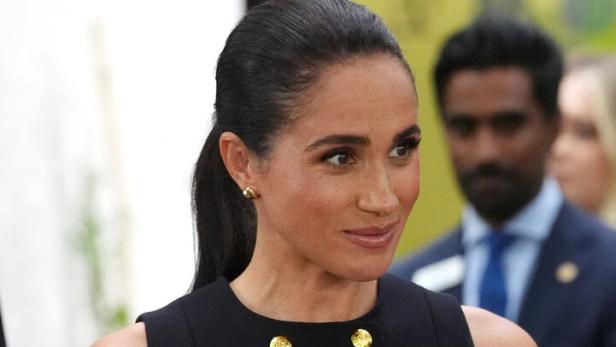 Meghan Markle trägt ein ärmelloses schwarzes Kleid mit goldenen Knöpfen und hat ihre Haare zu einem Pferdeschwanz zurückgebunden.