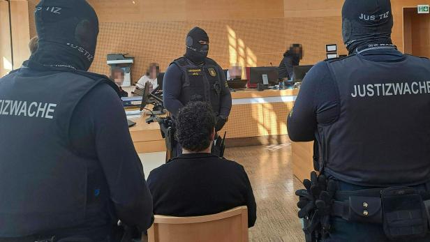 Mehrere Justizwachebeamte bewachen eine sitzende Person in einem Saal.