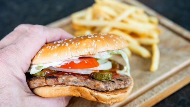 Eine Hand hält einen Burger mit Fleisch, Tomate und Gurken, während im Hintergrund Pommes frites auf einem Holzbrett liegen.