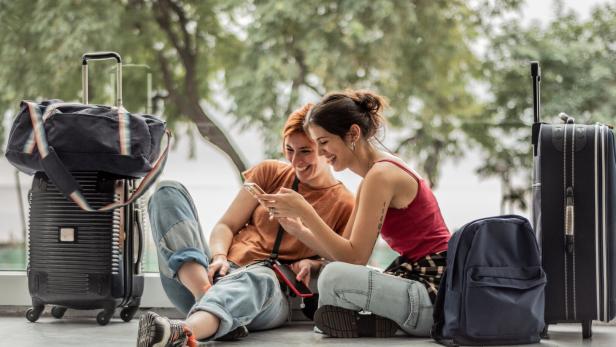 Zwei junge Frauen sitzen mit Koffern und Rucksäcken und lachen gemeinsam auf ein Smartphone.