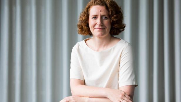 Eine Frau mit rötlichem, lockigem Haar und einem hellen Oberteil sitzt vor einem grauen, gestreiften Hintergrund.