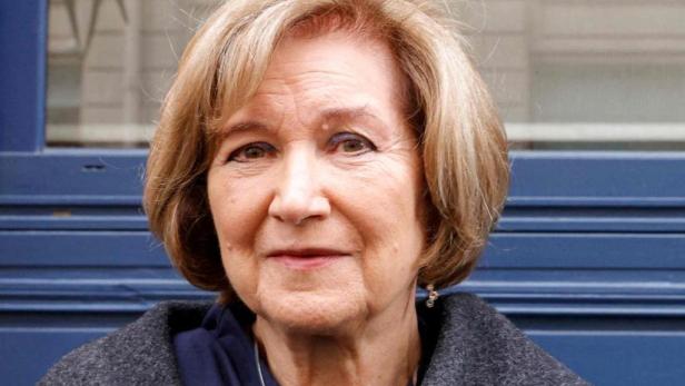 Eine ältere Frau mit kurzem, blondem Haar blickt freundlich vor einem blauen Hintergrund in die Kamera.