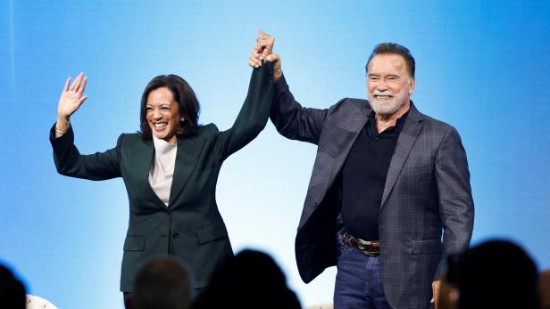Kamala Harris und Arnold Schwarzenegger stehen lächelnd vor einem blauen Hintergrund und halten gemeinsam die Hände in die Höhe.