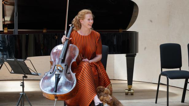 Eine lächelnde Cellistin in einem orange-schwarz gestreiften Kleid sitzt mit ihrem Instrument vor einem Flügel, während ein Hund zu ihren Füßen liegt.