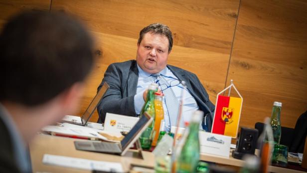 Mann im Anzug spricht an einem Konferenztisch vor Holzwand und Fahne.