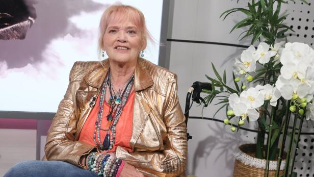 Gaby Köster mit blondem Haar trägt eine goldene Jacke, viele Ketten und Armbänder und sitzt in einem Rollstuhl neben einer weißen Orchidee.