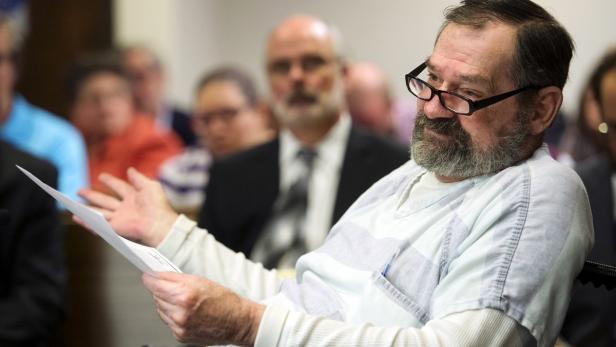 Ein Mann mit Brille und Bart gestikuliert mit einem Blatt Papier in der Hand vor Publikum.