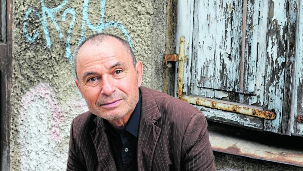 Ein Mann im braunen Nadelstreifenanzug lehnt vor einer verwitterten, graublauen Holztür mit Graffiti.
