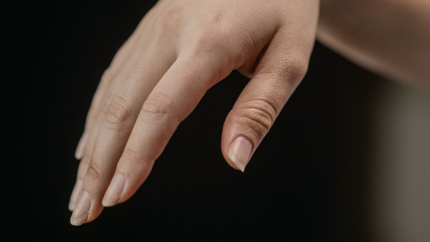 Nahaufnahme einer Hand mit brüchigen, teils gesplitteten Fingernägeln vor dunklem Hintergrund.