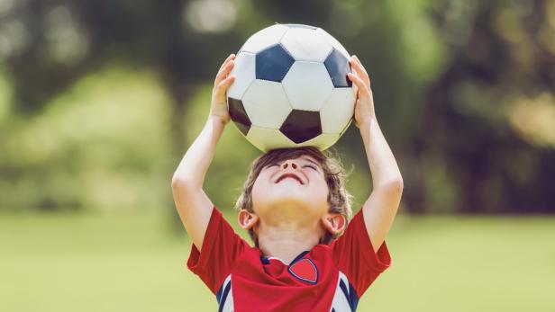 Ein Junge im roten Trikot hält einen Fußball über dem Kopf im Freien.