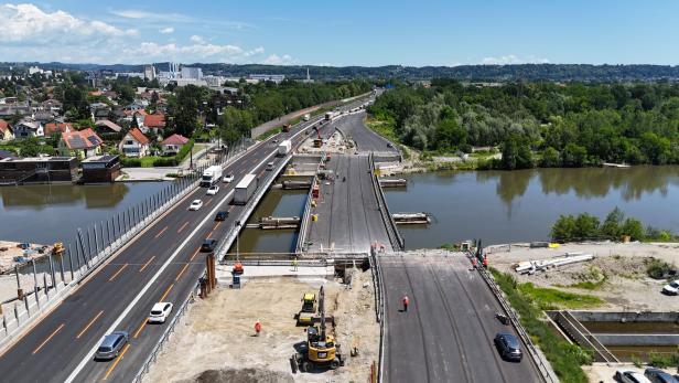 Luftaufnahme einer Brückenbaustelle über einen Fluss mit Autos, Baufahrzeugen und Stadt im Hintergrund.