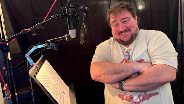 Ein lächelnder Mann steht vor Mikrofonen und einem Notenständer in einem Tonstudio.