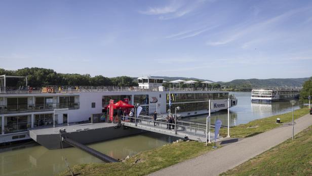 Anlegestelle am Fluss mit zwei Kreuzfahrtschiffen, Steg und Passagieren vor grünen Hügeln unter blauem Himmel.