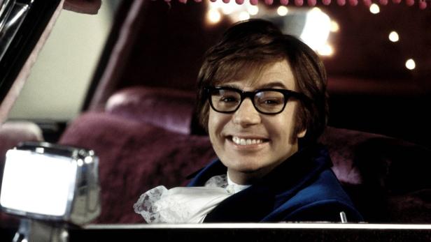 Mike Myers lächelt als Austin Powers mit Brille und blauem Samtanzug in einem Auto.