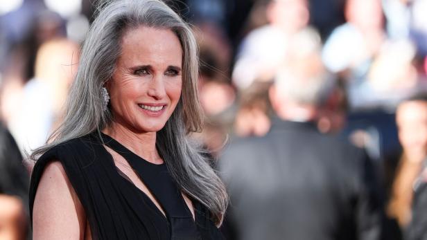 Andie MacDowell lächelt mit langem, grauem Haar in einem schwarzen Kleid vor einem unscharfen Hintergrund.