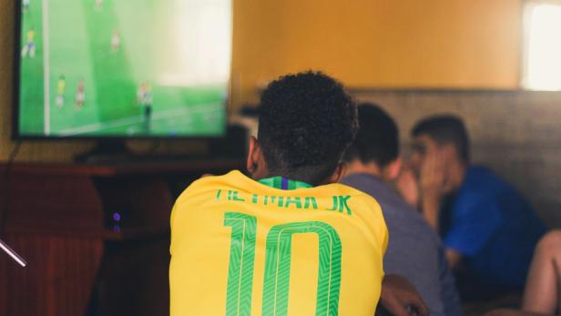 Ein Mann im Brasilien-Trikot Nummer 10 schaut mit anderen Zuschauern auf einen laufenden Fußballfernseher.
