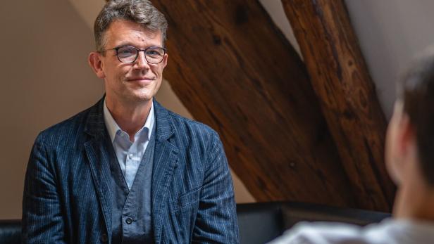 Ein Mann mit Brille und blauem Sakko sitzt in einem Gesprächsraum und blickt sein Gegenüber an.