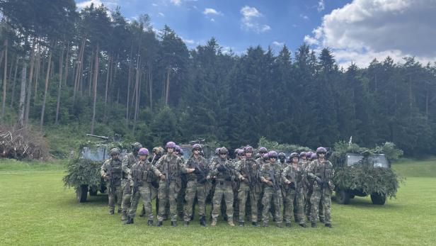 Soldaten mit Waffen posieren vor getarnten Militärfahrzeugen auf einer Wiese.