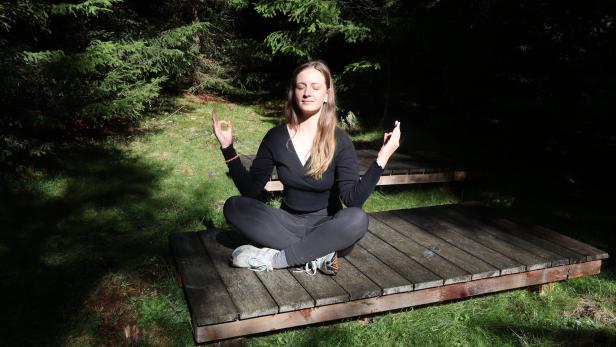 Frau meditiert im Lotussitz auf einer Holzplattform im Wald.