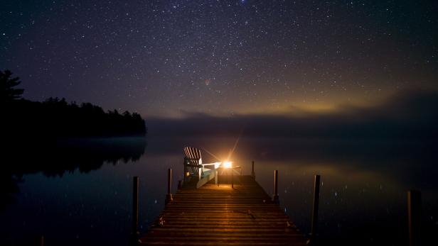 Sternenhimmel über einem stillen See, davor ein beleuchteter Holzsteg mit Stuhl.